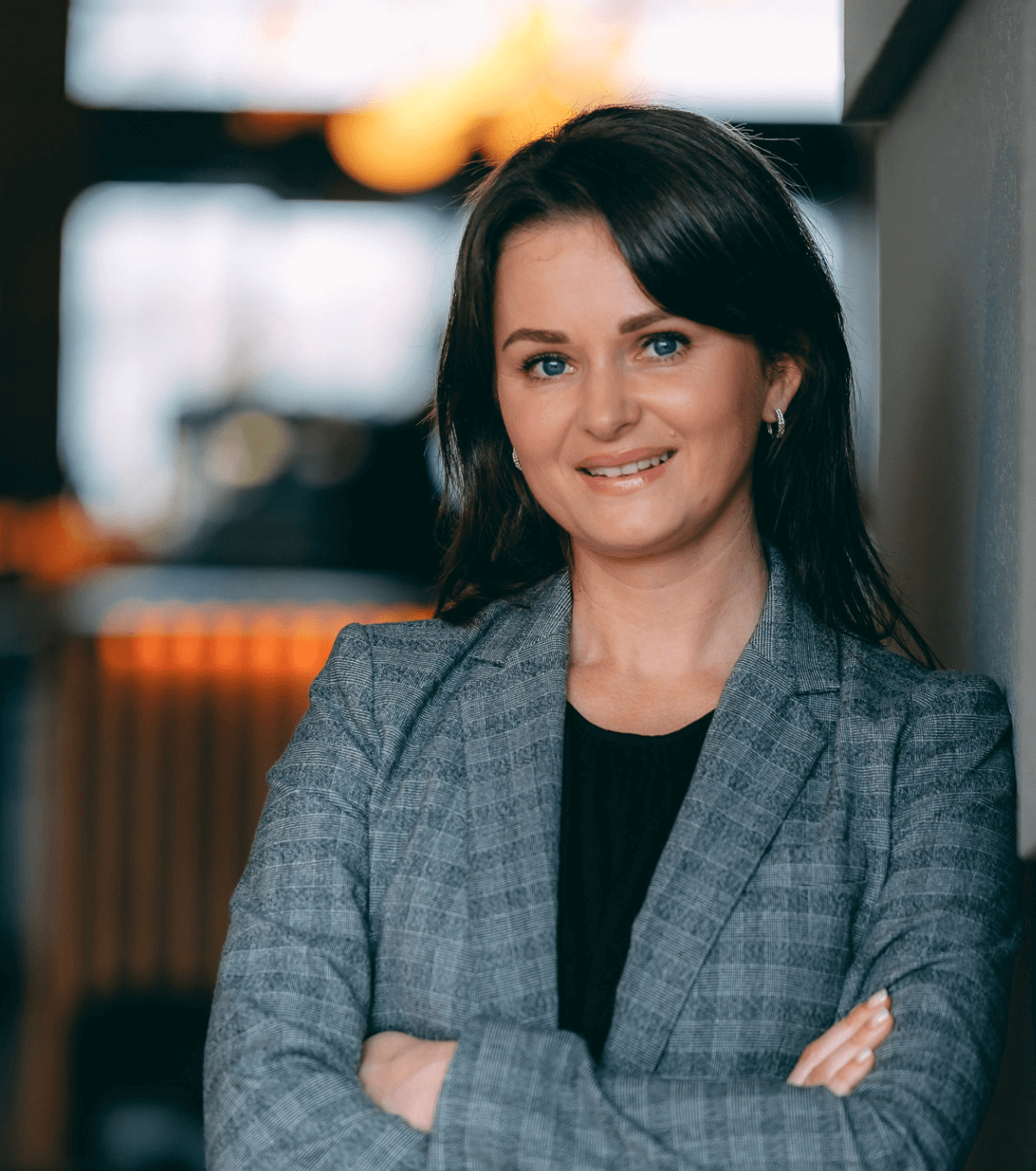 Profesjonalna kobieta z brązowymi włosami do ramion, ubrana w szarą marynarkę narzuconą na czarny top, uśmiecha się pewnie ze skrzyżowanymi ramionami. stoi w ciepło oświetlonym biurze.