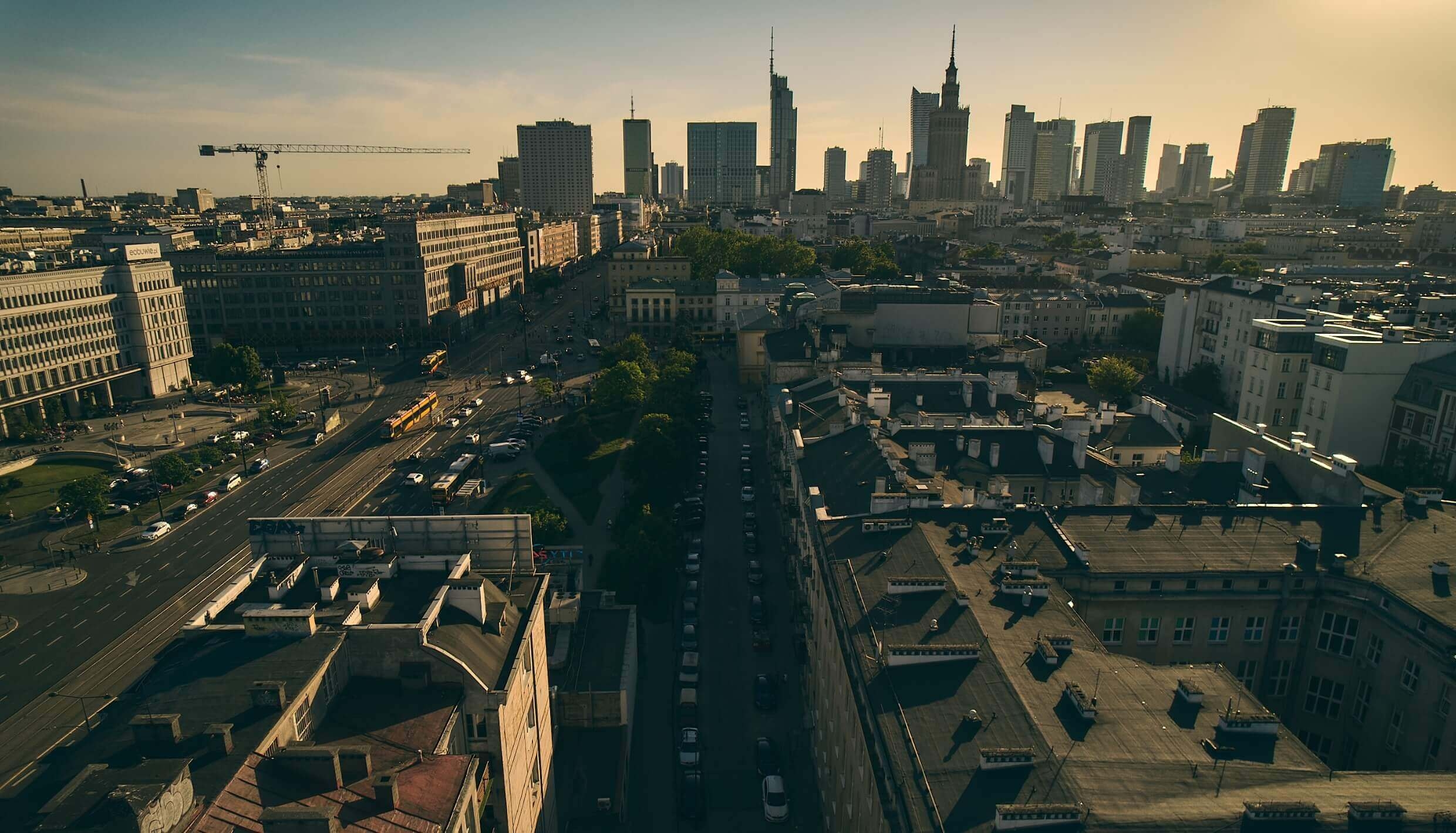 Widok z lotu ptaka na panoramę miasta o zachodzie słońca, z wysokimi nowoczesnymi wieżowcami w tle i starszymi budynkami na pierwszym planie. Ruchliwa ulica z samochodami i zielenią dzieli obszar miejski. Niebo jest czyste i ma ciepły, złoty odcień.