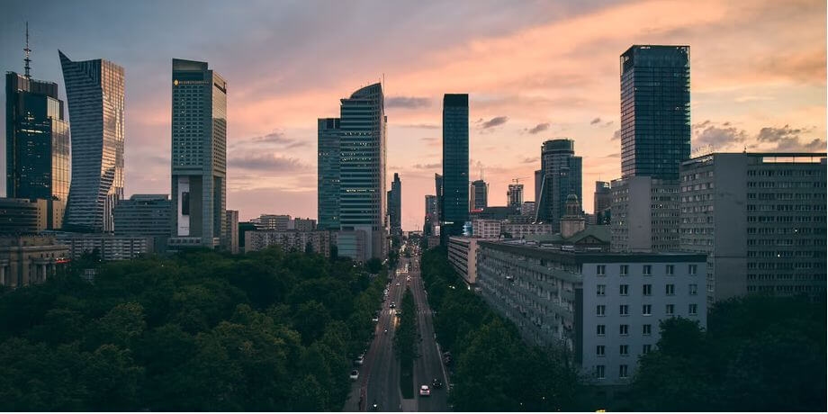 Panorama miasta o zachodzie słońca z drogą prowadzącą w dal. Wieżowce i nowoczesne budynki rysują się na tle kolorowego nieba w odcieniach pomarańczu, różu i błękitu. Drzewa na pierwszym planie, a samochody są widoczne na drodze.