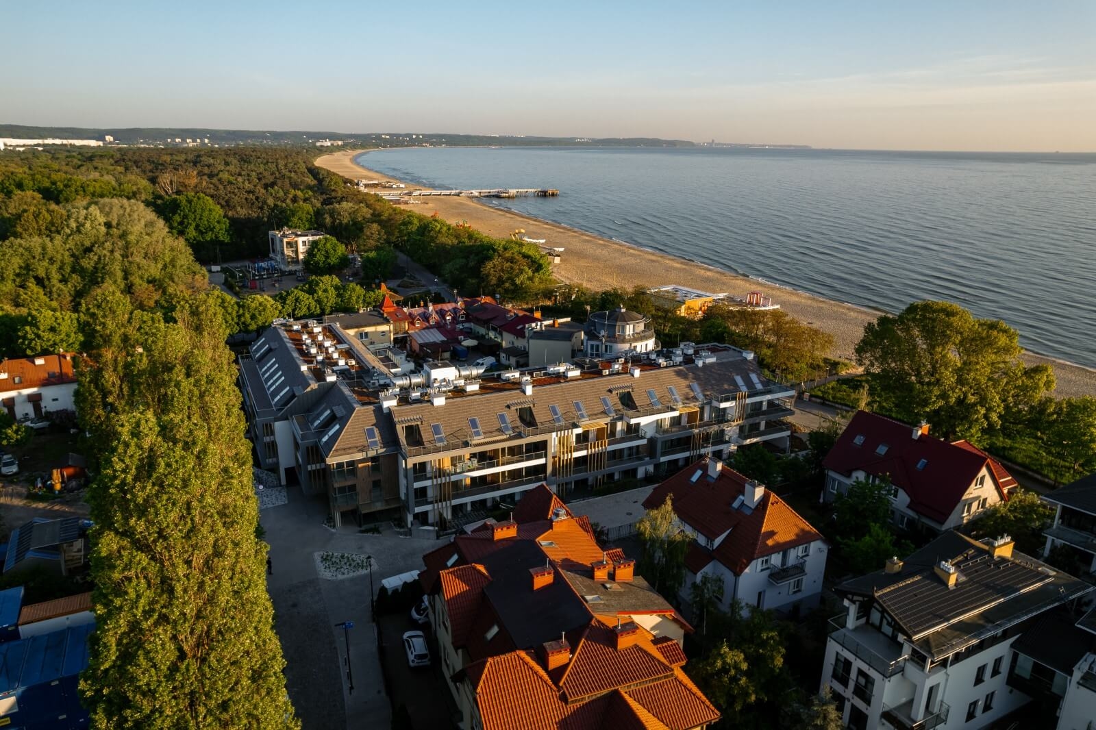 Widok z lotu ptaka na nadmorskie miasteczko z nowoczesnymi i tradycyjnymi budynkami z czerwonymi dachami. Linia brzegowa rozciąga się wzdłuż piaszczystej plaży otoczonej drzewami, pod czystym, błękitnym niebem i łagodnie toczącymi się falami morskimi.