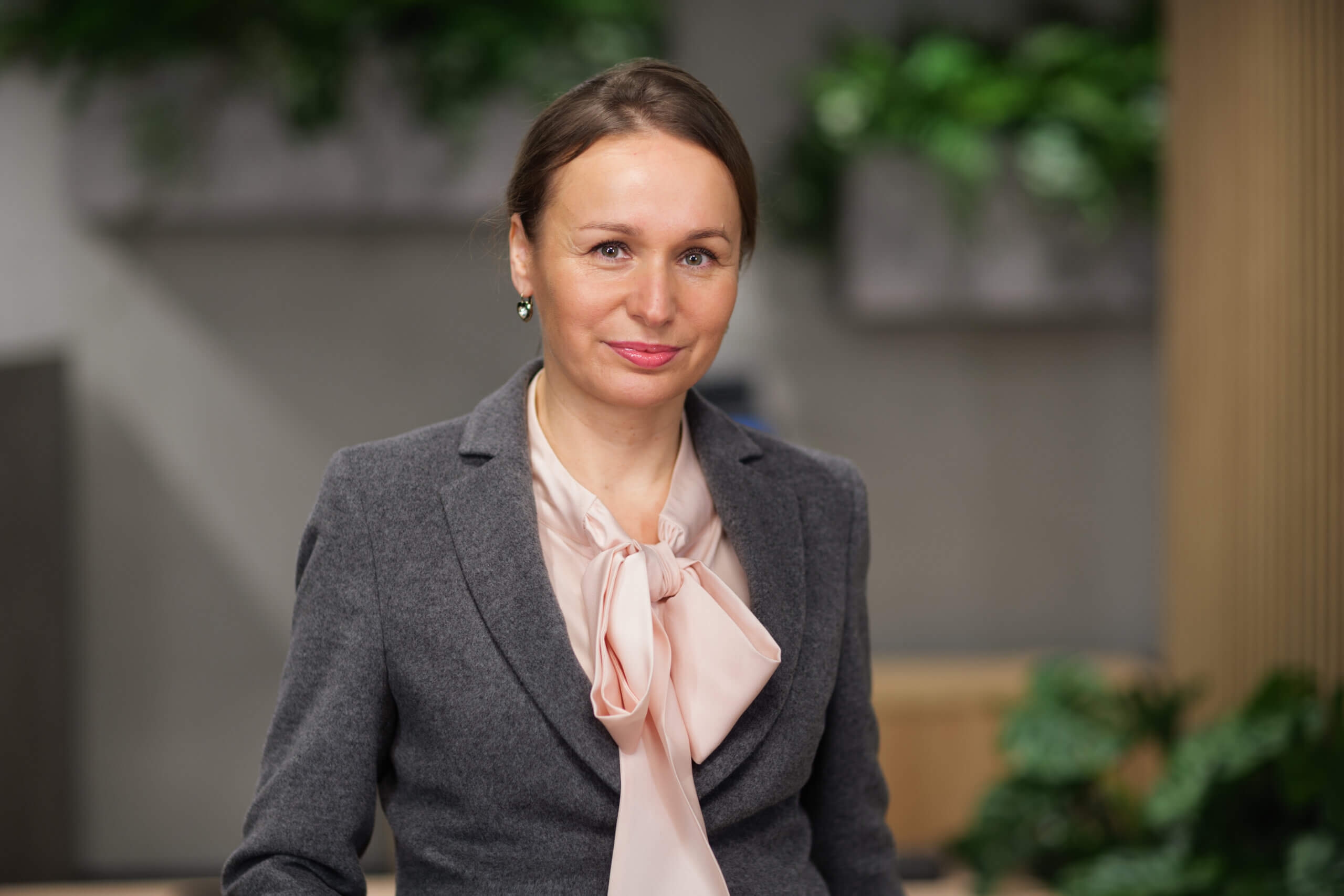 Kobieta w szarym garniturze z jasnoróżową bluzką stoi w środku. Ma jasnobrązowe włosy związane do tyłu i delikatnie się uśmiecha. Tło jest rozmyte, widać zielone rośliny.