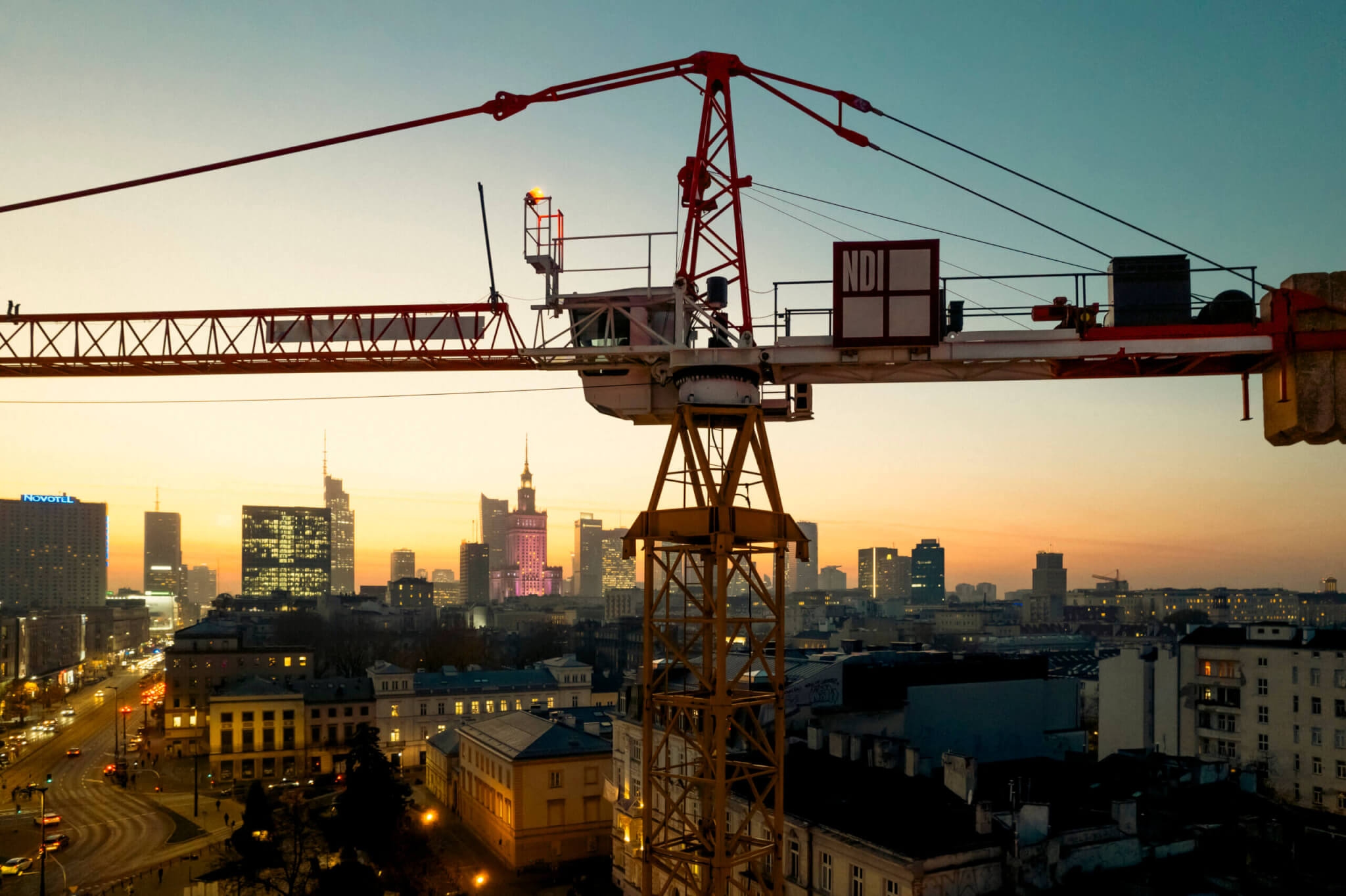 Duży dźwig budowlany unosi się nad miastem o zachodzie słońca. Na horyzoncie widać nowoczesne wieżowce i oświetlone budynki na tle nieba o zmierzchu, a ulice poniżej zaczynają się rozświetlać.