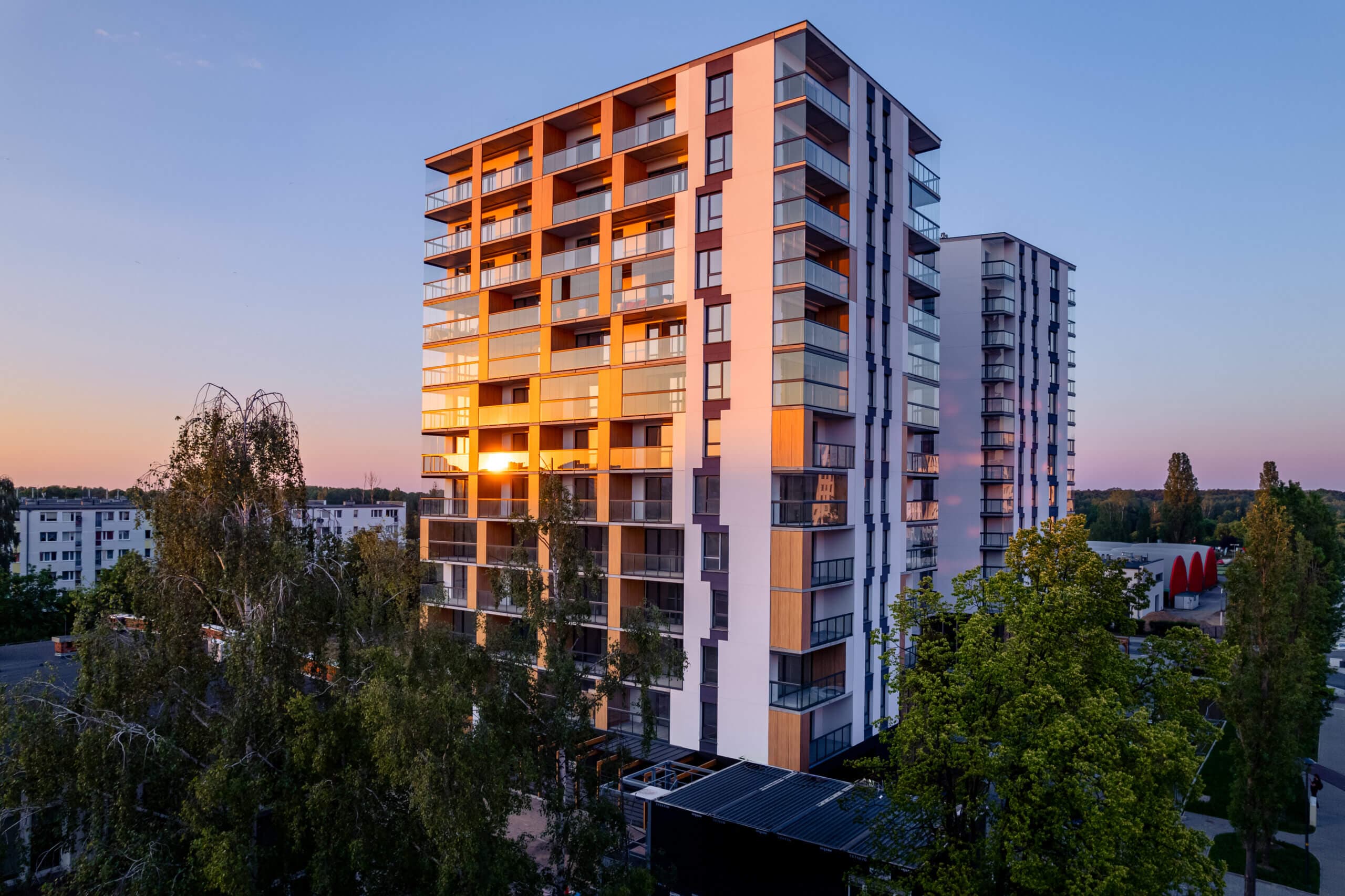 Nowoczesny wielopiętrowy budynek mieszkalny ze szklaną fasadą odbijającą zachód słońca. Budynek otoczony jest drzewami i innymi apartamentowcami. Scena została uchwycona wczesnym wieczorem, na tle czystego nieba.