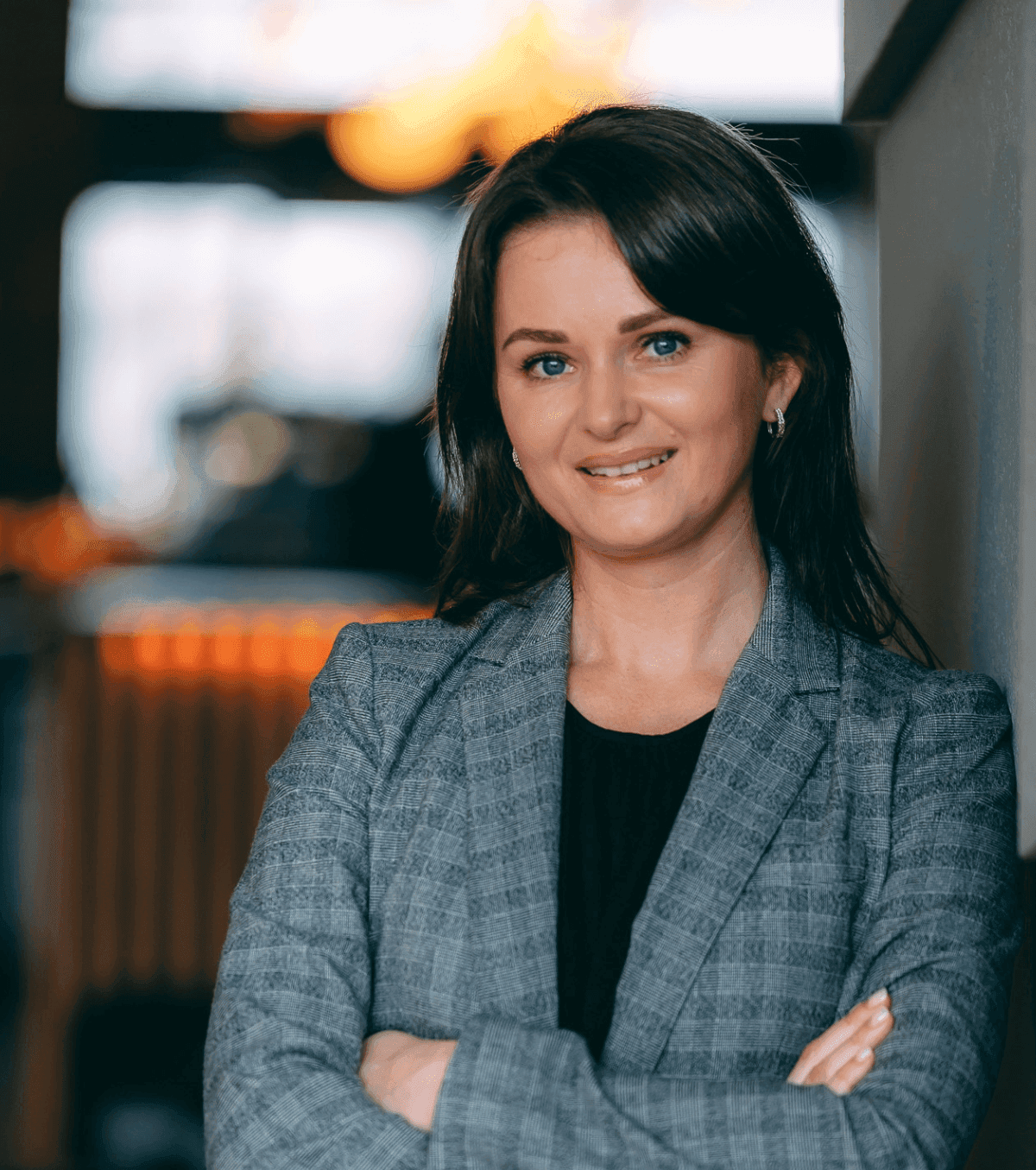 Profesjonalna kobieta z brązowymi włosami do ramion, ubrana w szarą marynarkę narzuconą na czarny top, uśmiecha się pewnie ze skrzyżowanymi ramionami. stoi w ciepło oświetlonym biurze.