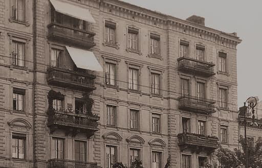 Czarno-białe zdjęcie zabytkowego, wielopiętrowego, ozdobnego budynku z balkonami ozdobionymi roślinami i markizami. Architektura obejmuje dekoracyjne kamienne elementy i kilka okien.