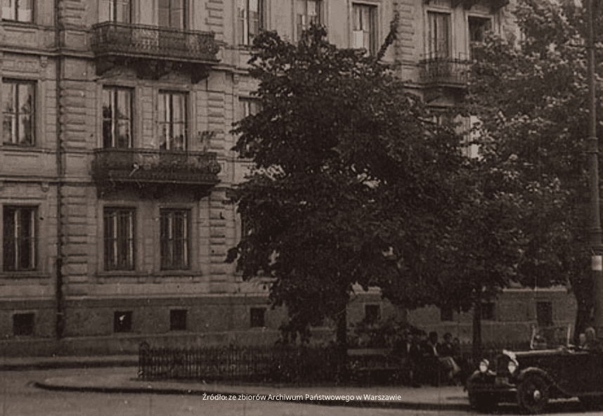 Czarno-białe zdjęcie zabytkowego budynku z ozdobnymi balkonami. Z przodu, zacienione drzewo stoi obok klasycznego samochodu zaparkowanego na rogu ulicy. Kilka osób siedzi na ławce w pobliżu.