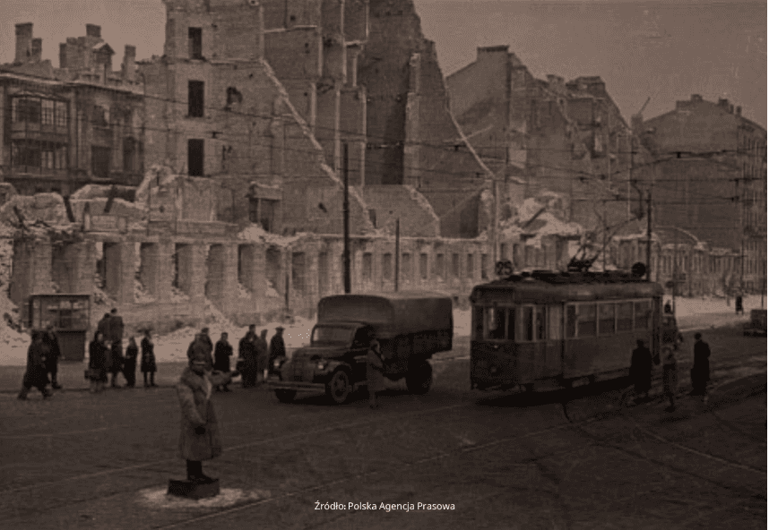 Historyczne czarno-białe zdjęcie przedstawia scenę uliczną z tramwajem i ciężarówką przed zniszczonymi przez wojnę budynkami. Ludzie zgromadzili się na chodniku, a nad nimi widać linie energetyczne.
