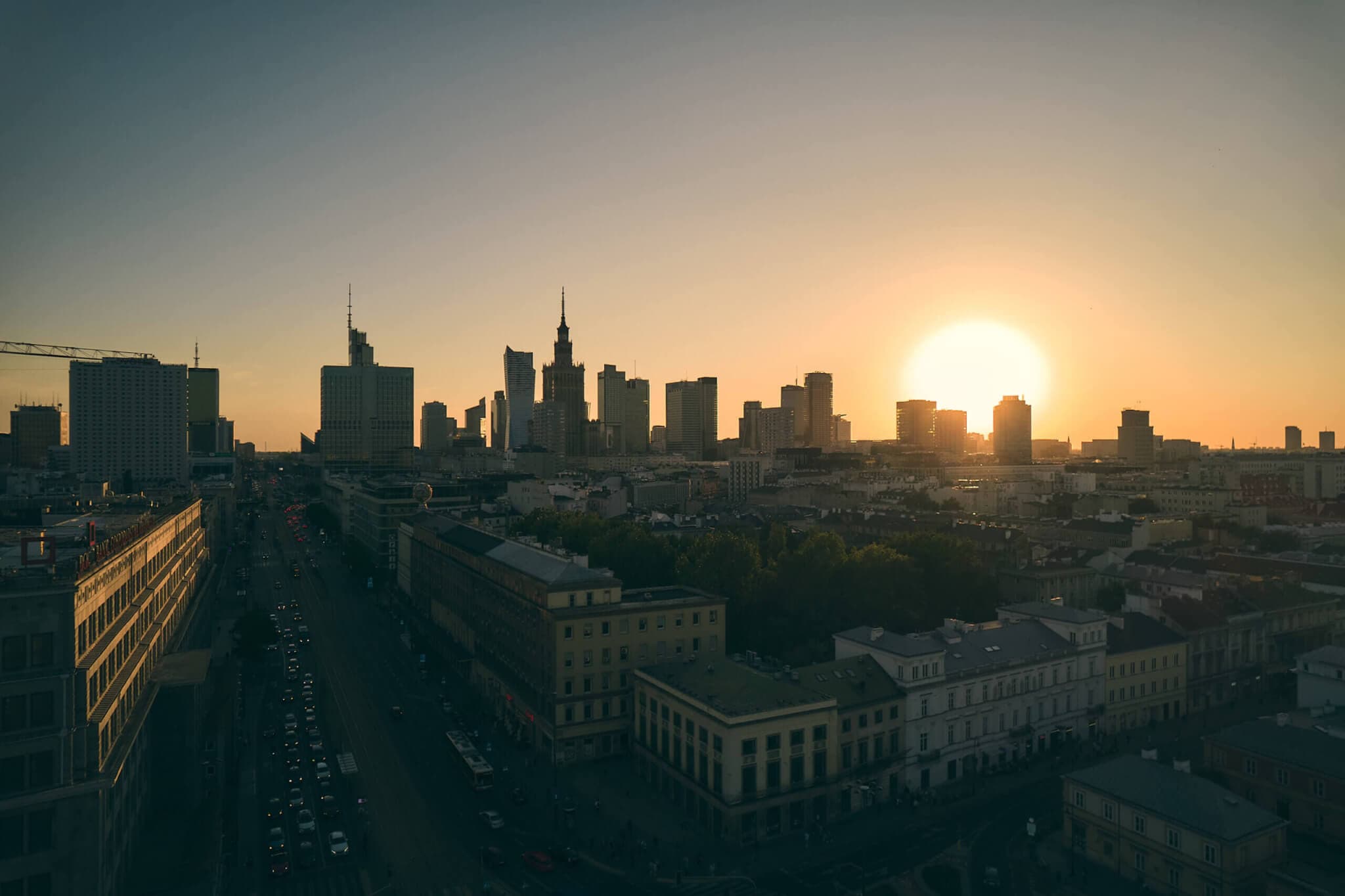 Krajobraz miejski o zachodzie słońca, z zachodzącym słońcem za nowoczesną linią wysokich budynków. Ulica z rzadkim ruchem ulicznym przebiega przez pierwszy plan, a ciepły blask zachodzącego słońca rzuca długie cienie i pomarańczowe odcienie na całą scenę.