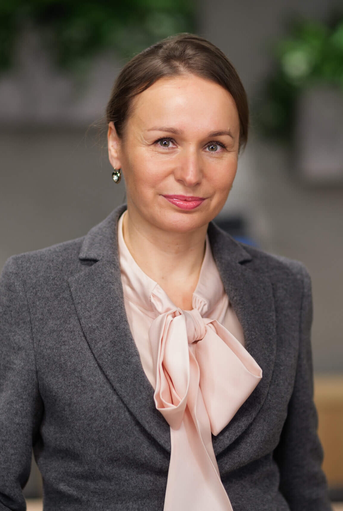 Kobieta w szarym garniturze z jasnoróżową bluzką stoi w środku. Ma jasnobrązowe włosy związane do tyłu i delikatnie się uśmiecha. Tło jest rozmyte, widać zielone rośliny.